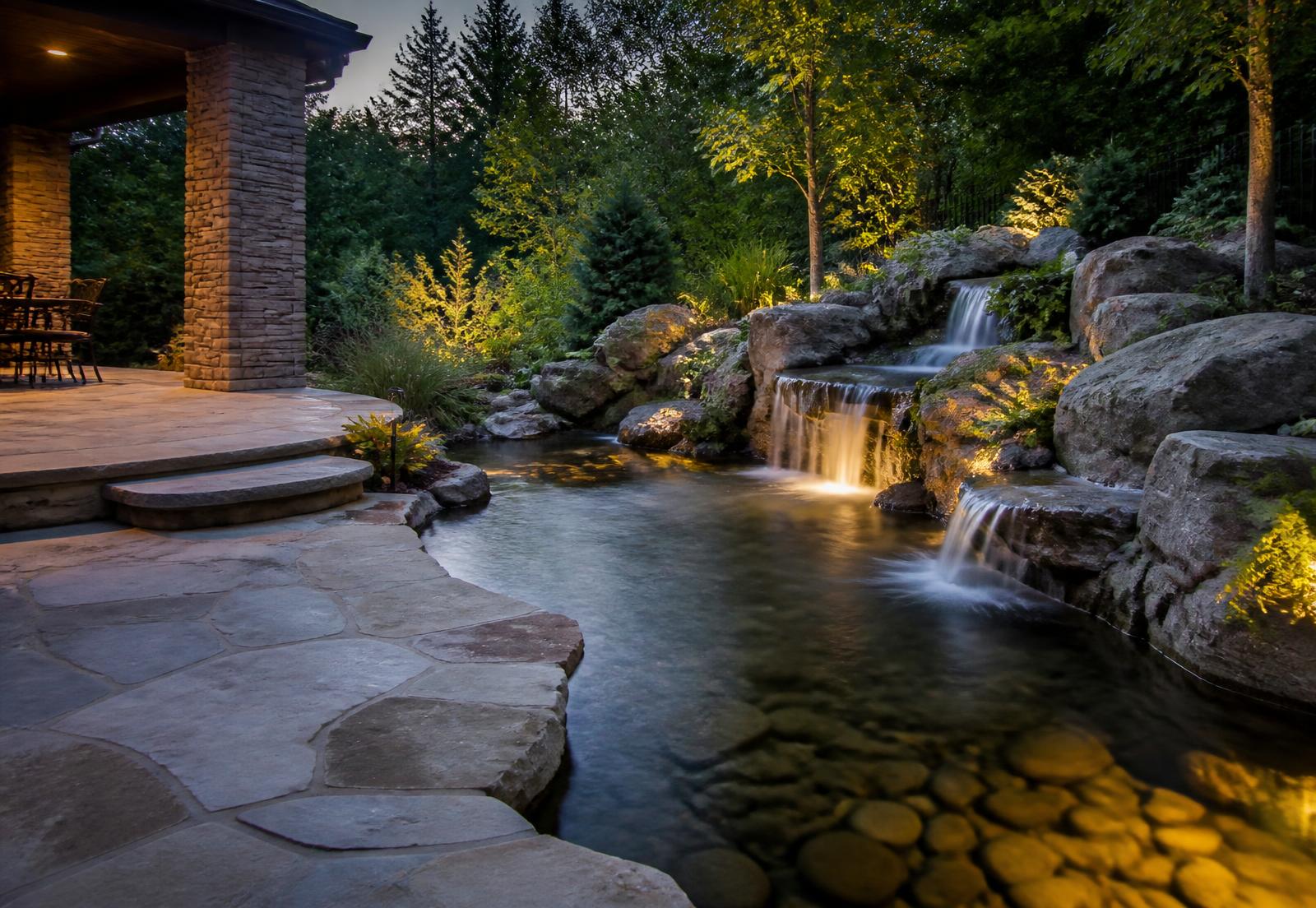 Stone patio hardscape with a rock waterfall flowing into a landscaped pond — representative imagery